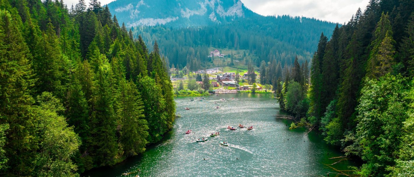 Aerial drone view of Red Lake in Romania. Lake in a valley in Carpathian mountains, multiple people floating on boats, lush forest around, village in the distance